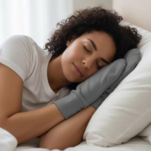May include: A person sleeping on a white pillow, wearing a gray sleep mitt. The person is wearing a white t-shirt. The mitt covers the hand and wrist, and is made of a soft, gray fabric.