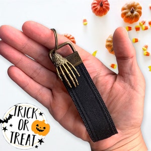 May include: Black fabric keychain with a skeleton hand charm. The keychain has a zig-zag pattern and the text "trick or treat" is on a white circle with a black bat and orange pumpkin.
