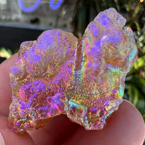 Puede incluir: Cristal iridiscente con una superficie texturizada, que muestra una gama de colores que incluyen rosa, morado, amarillo y verde. La palabra "Crystals" es visible en el fondo.