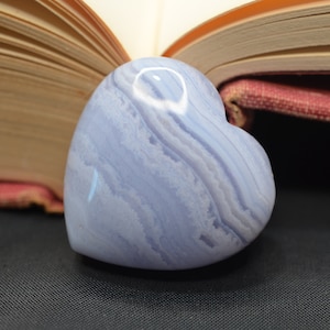 May include: A heart-shaped blue agate stone with white and gray banding. The stone is resting on a closed book with a red cover.