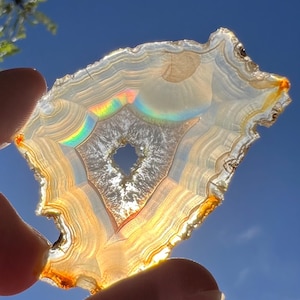 May include: A translucent agate slice, held against a bright blue sky. The stone features concentric bands of cream, brown, and orange, with a central cavity. A rainbow effect is visible within the stone, adding a colorful accent to the natural pattern.