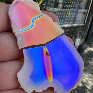 May include: Two iridescent, fractured pieces of a stone, held in a hand. The top piece is pink and orange with a blue line. The bottom piece is blue with a rainbow streak. The stone has a natural, rough edge.
