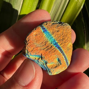 May include: A small, iridescent, orange-brown stone with blue and green hues. The stone has a rough, textured surface.