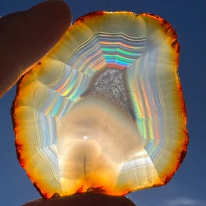 May include: A colourful agate slice, displaying bands of white, orange, and red. The stone has a central area with a rainbow effect. The agate is held against a blue sky, showing its natural beauty. The agate's measurements are in centimetres.