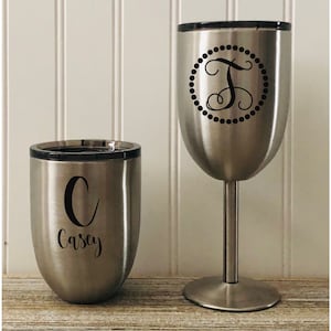 May include: Two stainless steel tumblers with black lids. The tumbler on the left has a black letter "C" and the name "Casey" printed on it. The tumbler on the right has a black letter "T" inside a circle with black dots around it.
