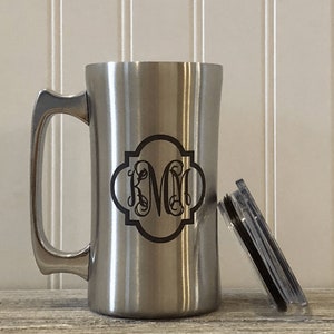 Puede incluir: Una taza de viaje de acero inoxidable con un dise&ntilde;o de monograma negro. La taza tiene un asa y una tapa.