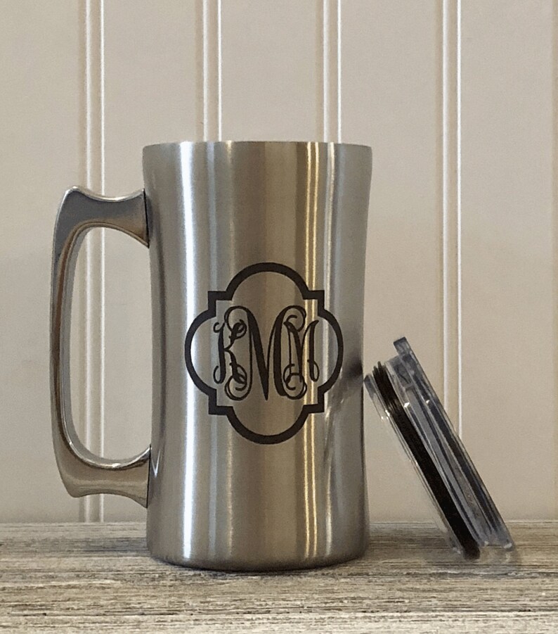Puede incluir: Una taza de viaje de acero inoxidable con un dise&ntilde;o de monograma negro. La taza tiene un asa y una tapa.