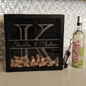 May include: A black wooden box with a glass front, featuring a custom engraved design with the names "Shwetha & Andrew" and the date "04.14.18". The box is filled with wine corks and is a unique way to commemorate special occasions.