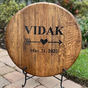 Op de afbeelding: Een rond houten bord met de tekst "VIDAK" en een pijl met een hart in het midden. De datum "May 21, 2022" staat onder de pijl.