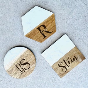 Puede incluir: Tres posavasos redondos de madera con una parte superior de mármol blanco. Los posavasos tienen los nombres "Ross", "Stein" y "Mr. & Mrs." grabados en ellos.