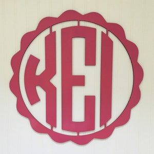Puede incluir: Un monograma de madera rosa con las letras "KEI" en blanco, recortadas en un círculo con un borde festoneado.