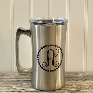 Puede incluir: Una taza de viaje de acero inoxidable con una tapa negra y un monograma "A" en un c&iacute;rculo. La taza tiene un asa y est&aacute; dise&ntilde;ada para bebidas calientes o fr&iacute;as.