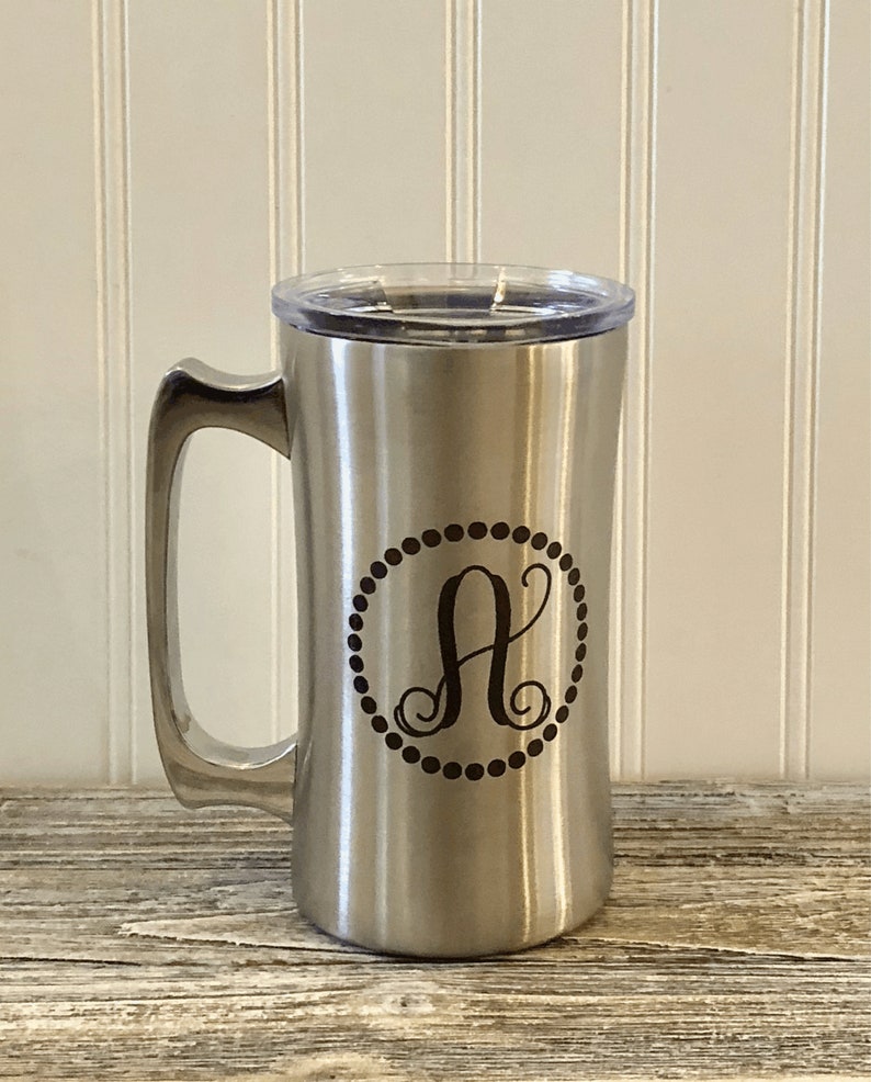 Puede incluir: Una taza de viaje de acero inoxidable con una tapa negra y un monograma "A" en un c&iacute;rculo. La taza tiene un asa y est&aacute; dise&ntilde;ada para bebidas calientes o fr&iacute;as.