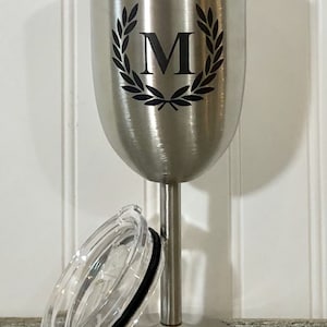 May include: Stainless steel wine tumbler with a lid. The tumbler has a black and white design of a laurel wreath with the letter "M" in the center.