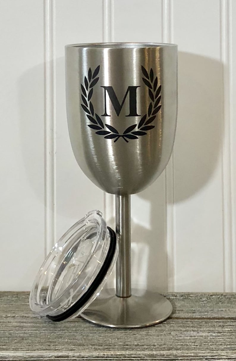 May include: Stainless steel wine tumbler with a lid. The tumbler has a black and white design of a laurel wreath with the letter "M" in the center.