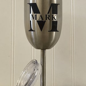 May include: A stainless steel wine tumbler with a black vinyl decal that says "M MARK IV". The tumbler has a clear lid.