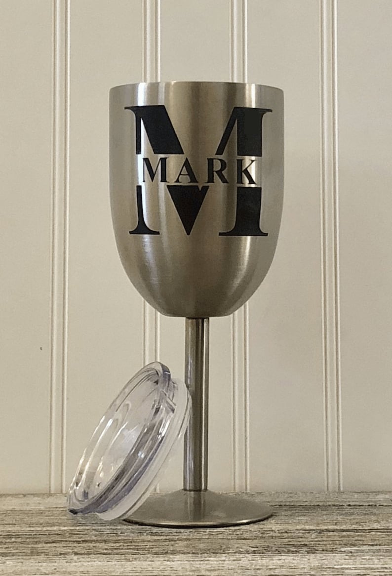 May include: A stainless steel wine tumbler with a black vinyl decal that says "M MARK IV". The tumbler has a clear lid.