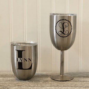 May include: Two stainless steel tumblers with lids. The tumbler on the left has the letter L and the name Lynn printed on it. The tumbler on the right has a monogram of the letter L inside a circle.