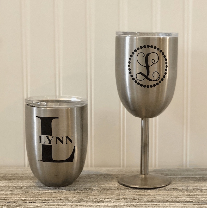 May include: Two stainless steel tumblers with lids. The tumbler on the left has the letter L and the name Lynn printed on it. The tumbler on the right has a monogram of the letter L inside a circle.