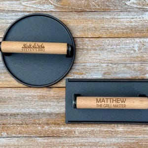 May include: Two black cast iron grill press with wooden handles. The top handle has the text "STEVEN'S BBQ" and flames. The bottom handle has the text "MATTHEW THE GRILL MASTER".