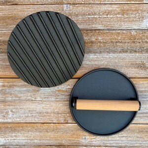 May include: Two black cast iron round cooking presses. One press has a ridged surface and the other has a smooth surface with a wooden handle.
