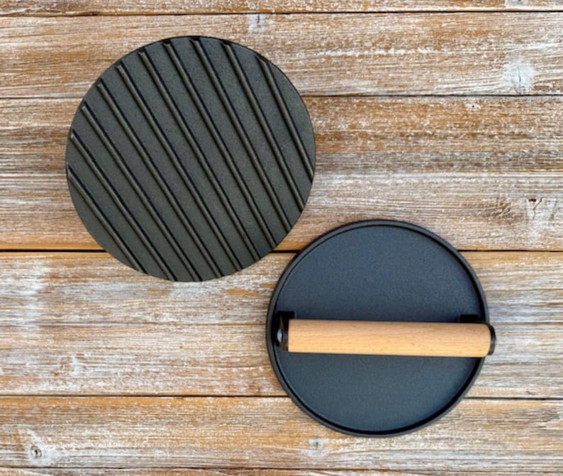 May include: Two black cast iron round cooking presses. One press has a ridged surface and the other has a smooth surface with a wooden handle.