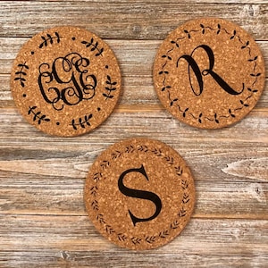 May include: Three round cork trivets with black engraved monograms. The trivets feature a decorative leaf border. The monograms are "GG", "R", and "S".