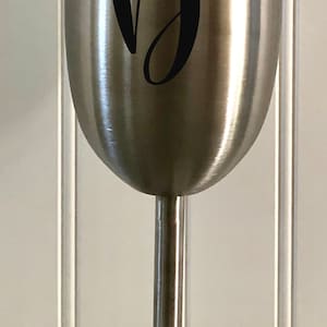 May include: A stainless steel wine tumbler with a black letter "D" on the side. The tumbler has a lid and a stem.