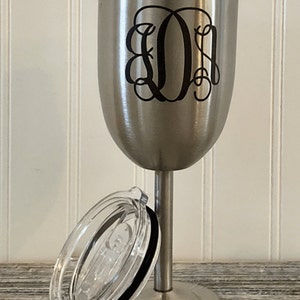 May include: A stainless steel wine tumbler with a black monogrammed design and a clear lid. The tumbler has a stem and a wide base.