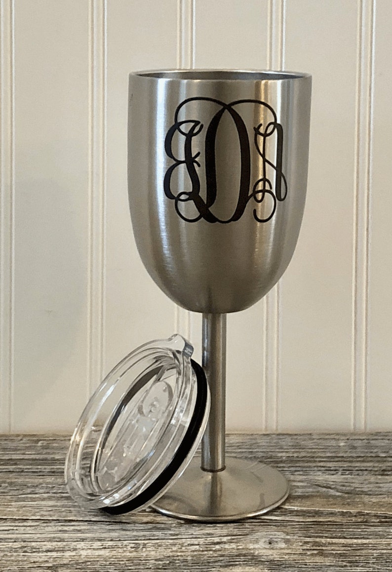 May include: A stainless steel wine tumbler with a black monogrammed design and a clear lid. The tumbler has a stem and a wide base.