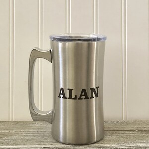 Puede incluir: Una taza de viaje de acero inoxidable con tapa y asa. La taza tiene el nombre "ALAN" grabado en la parte delantera.