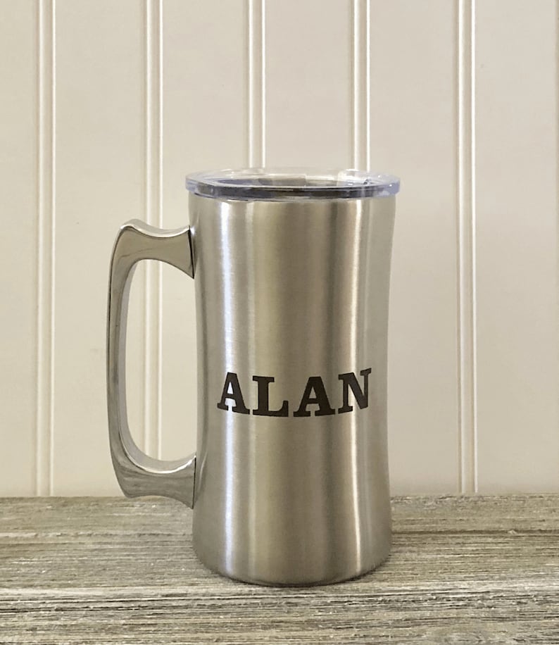 Puede incluir: Una taza de viaje de acero inoxidable con tapa y asa. La taza tiene el nombre "ALAN" grabado en la parte delantera.