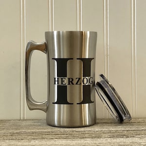 Puede incluir: Una taza de viaje de acero inoxidable con un asa y una tapa negras. La taza tiene la letra "H" impresa en la parte delantera, con el nombre "HERZOG" impreso debajo de la letra.