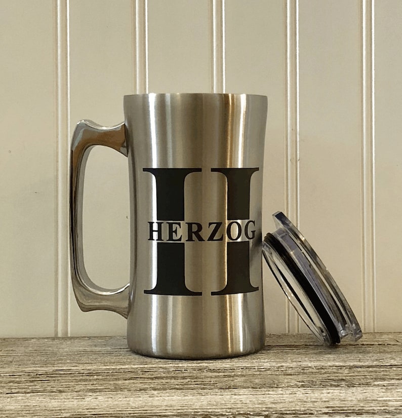 Puede incluir: Una taza de viaje de acero inoxidable con un asa y una tapa negras. La taza tiene la letra "H" impresa en la parte delantera, con el nombre "HERZOG" impreso debajo de la letra.