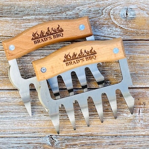 May include: Two wooden handled meat claws with a stainless steel claw design. The handles are engraved with "Brad's BBQ" and a flame design.
