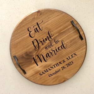 May include: A round wooden serving tray with black metal handles. The tray has the text "Eat Drink and be Married" and "Samantha & Alex October 28, 2023" engraved on it.