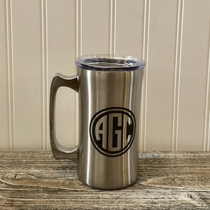 Puede incluir: Una taza de viaje de acero inoxidable con una tapa negra y un monograma negro de las letras "AGC" en un c&iacute;rculo en la parte delantera.