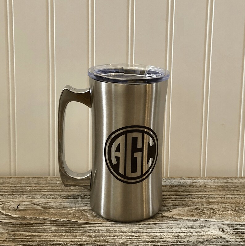 Puede incluir: Una taza de viaje de acero inoxidable con una tapa negra y un monograma negro de las letras "AGC" en un c&iacute;rculo en la parte delantera.