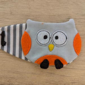 May include: A hair clip featuring a gray owl applique with large white eyes, black pupils, a yellow beak, and orange wings and belly. The owl is attached to a fabric base with a gray and white horizontal stripe pattern and a black metal clip.