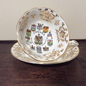 May include: A white teacup and saucer with gold trim and a design of the coats of arms of the Canadian provinces. The text "CANADA Coats of Arms & Emblems" is printed on the cup.