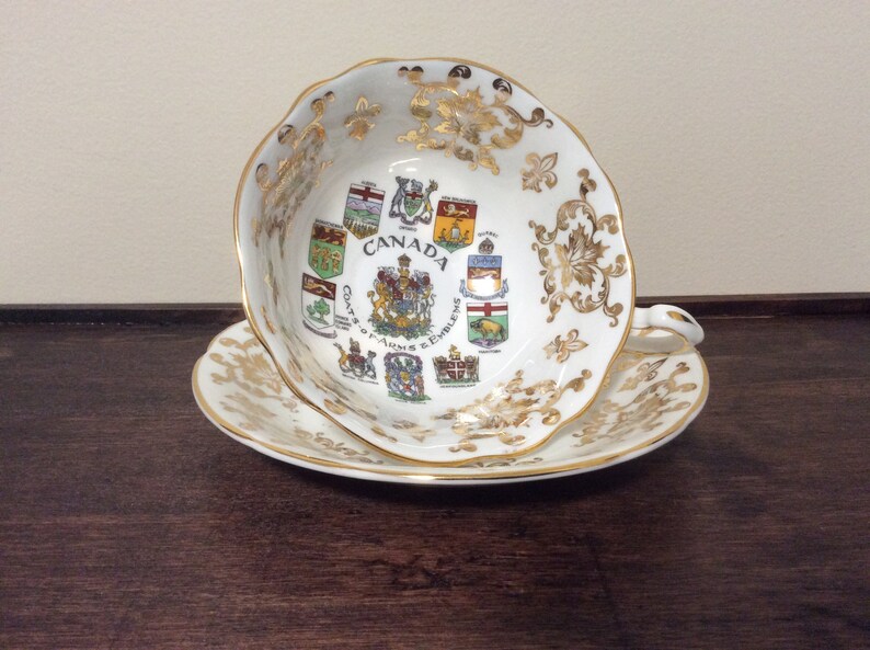May include: A white teacup and saucer with gold trim and a design of the coats of arms of the Canadian provinces. The text "CANADA Coats of Arms & Emblems" is printed on the cup.