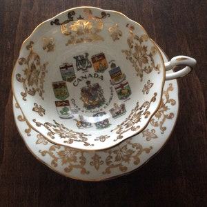 May include: A white teacup and saucer with gold trim and a design of the coats of arms of the provinces of Canada. The text "Canada Coats of Arms & Emblems" is printed on the cup.