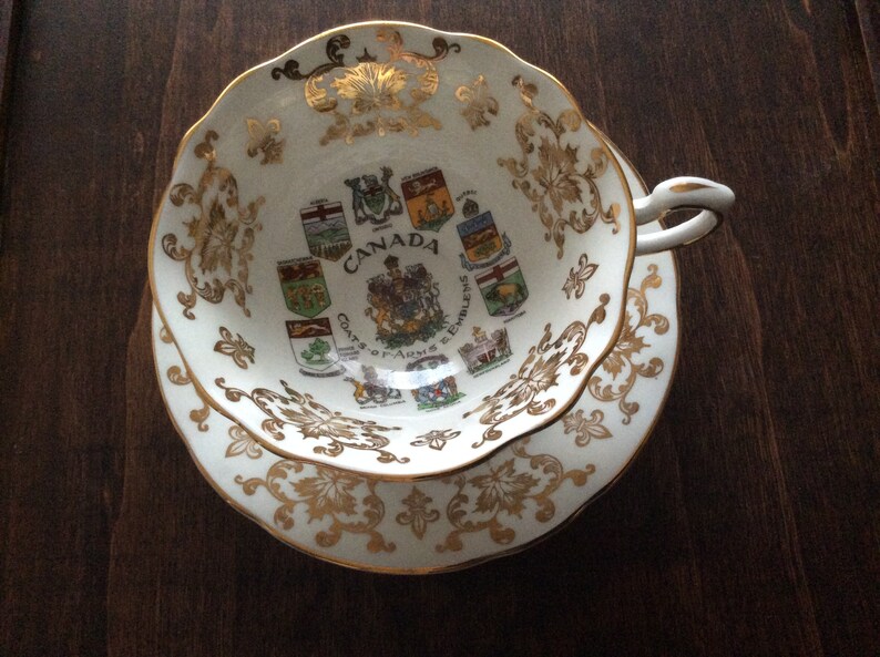 May include: A white teacup and saucer with gold trim and a design of the coats of arms of the provinces of Canada. The text "Canada Coats of Arms & Emblems" is printed on the cup.