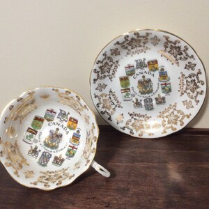May include: A white teacup and saucer with gold trim and a design of the coats of arms of the Canadian provinces. The text "CANADA" is printed in the center of the design.