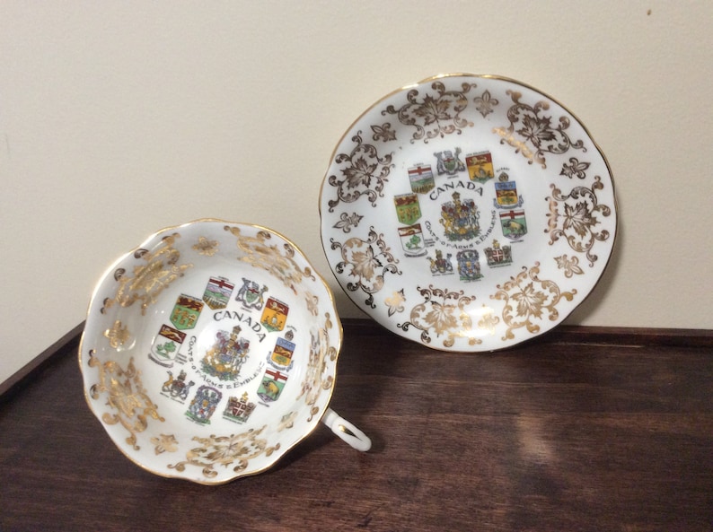 May include: A white teacup and saucer with gold trim and a design of the coats of arms of the Canadian provinces. The text "CANADA" is printed in the center of the design.