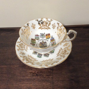 May include: A white teacup and saucer with gold trim and a design of the coats of arms of the Canadian provinces. The text "CANADA" is printed on the cup.