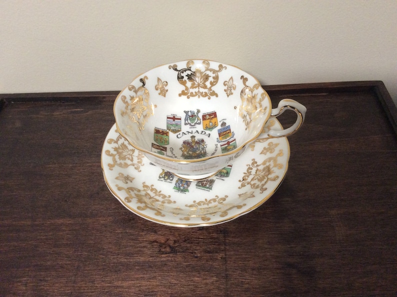 May include: A white teacup and saucer with gold trim and a design of the coats of arms of the Canadian provinces. The text "CANADA" is printed on the cup.