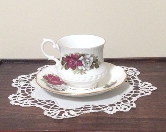 May include: A white teacup and saucer with a floral design featuring red and white roses. The cup and saucer have a gold trim.