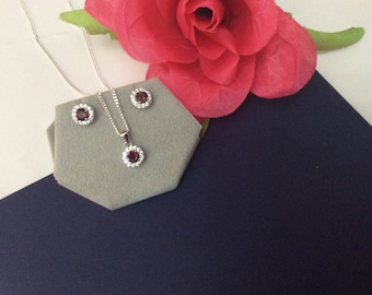 May include: A silver necklace and earrings set with a red gemstone surrounded by a halo of smaller white gemstones. The jewellery is displayed on a grey hexagonal cushion.