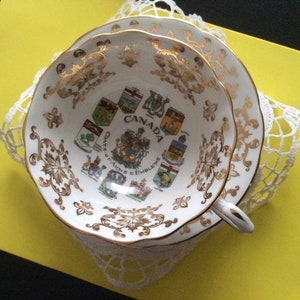 May include: A white teacup and saucer with gold trim and a design of the Canadian Coat of Arms and the provincial coats of arms. The text "CANADA" and "COATS OF ARMS & EMBLEMS" are printed on the cup.